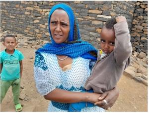 A mother holds her child close after receiving a vaccination, with another young sibling standing nearby. The family is pictured outside their home, illustrating the community's participation in vaccination efforts to protect children’s health.