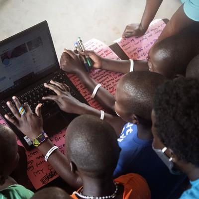 A group of children gather around a laptop, working together on a digital project, with handwritten notes and pens visible, highlighting youth engagement in digital education and teamwork.
