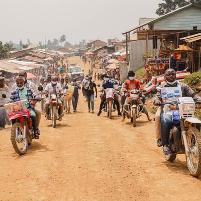 A group of vaccination workers on motorcycles ride through a rural town, wearing masks and vaccination campaign banners. The motorcycles navigate a dirt road lined with local shops and people. The workers are participating in a polio vaccination outreach effort.