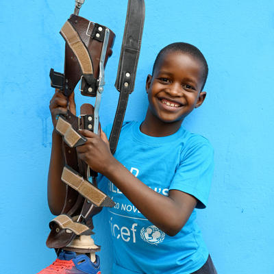 Smiling child in a UNICEF t-shirt holding an orthopedic leg brace against a bright blue background.