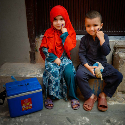 Two children smiling and showing vaccination marks, sitting next to a polio vaccine cold box.