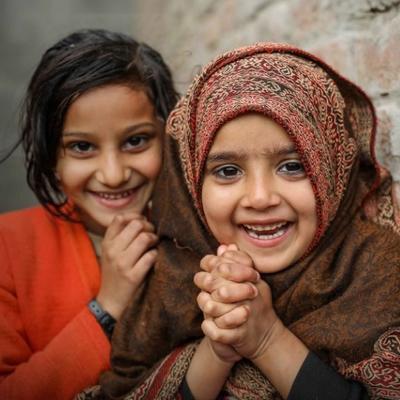 Two smiling girls, one in an orange sweater and the other in a patterned shawl, leaning against a brick wall.