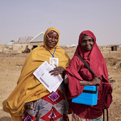 Two women wearing colorful hijabs stand in a dusty rural setting, holding a paper checklist and a small cooler used for medical supplies.
