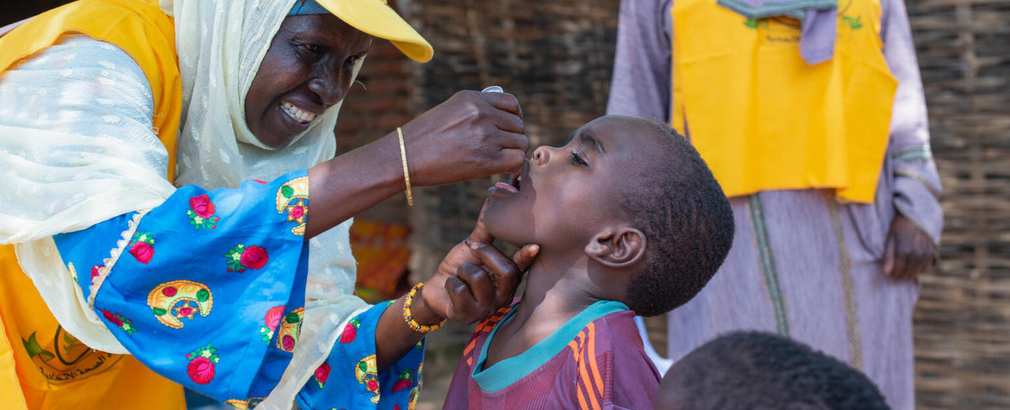 A smiling health worker in a yellow vest and cap administers oral vaccine drops to a young boy who tilts his head back with his mouth open, while another child stands nearby watching.