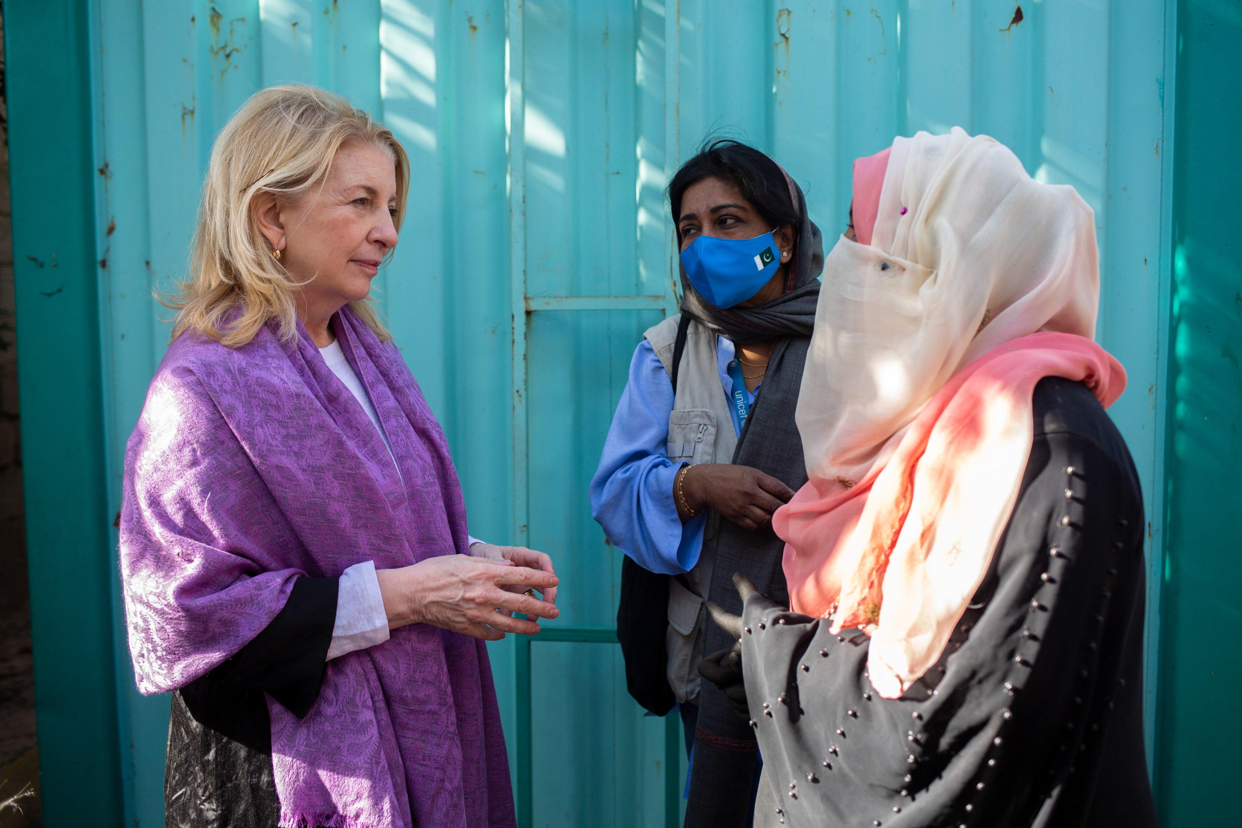 Catherine Russell, wearing a purple shawl, speaks with two women, one in a black and pink headscarf and another in a blue UNICEF vest and face mask, against a turquoise metal background.
