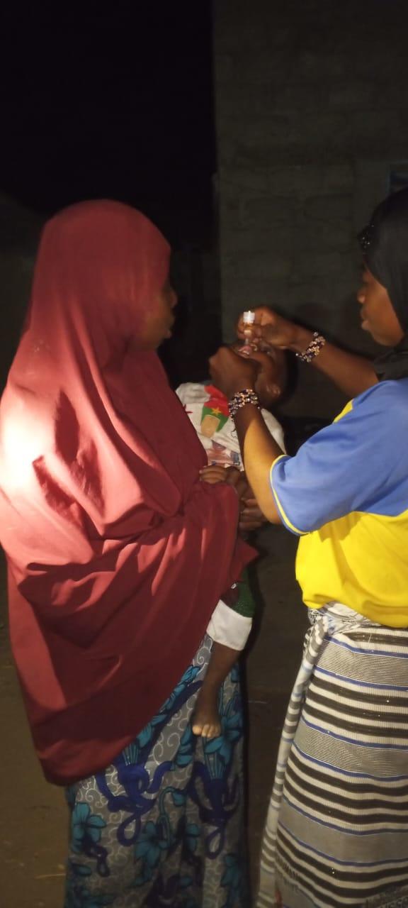 A female health worker in a yellow and blue uniform administers oral polio vaccine drops to a young child held by the mother wearing a pink/red hijab. Another woman stands watching. The scene takes place at night, with darkness visible in the background.