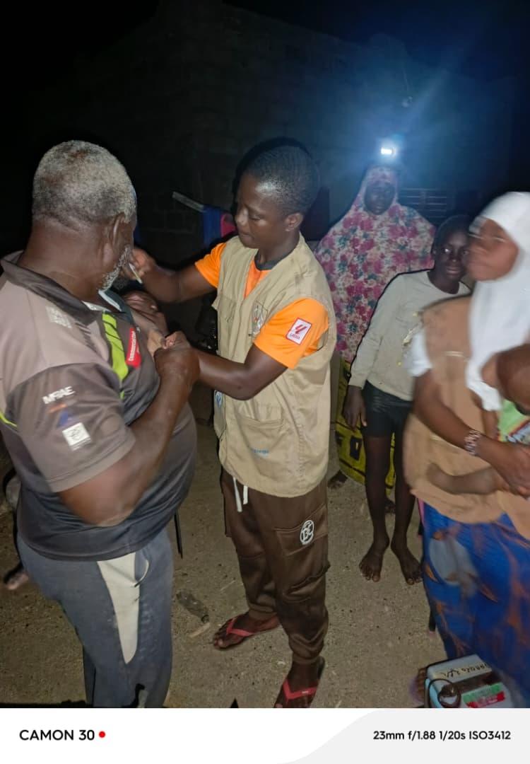 A male health worker wearing a beige vest over an orange shirt vaccinates an older man's arm at night in Zabré. Other community members, including women in traditional dress, watch the vaccination. A bright light source illuminates the scene against the dark night background.