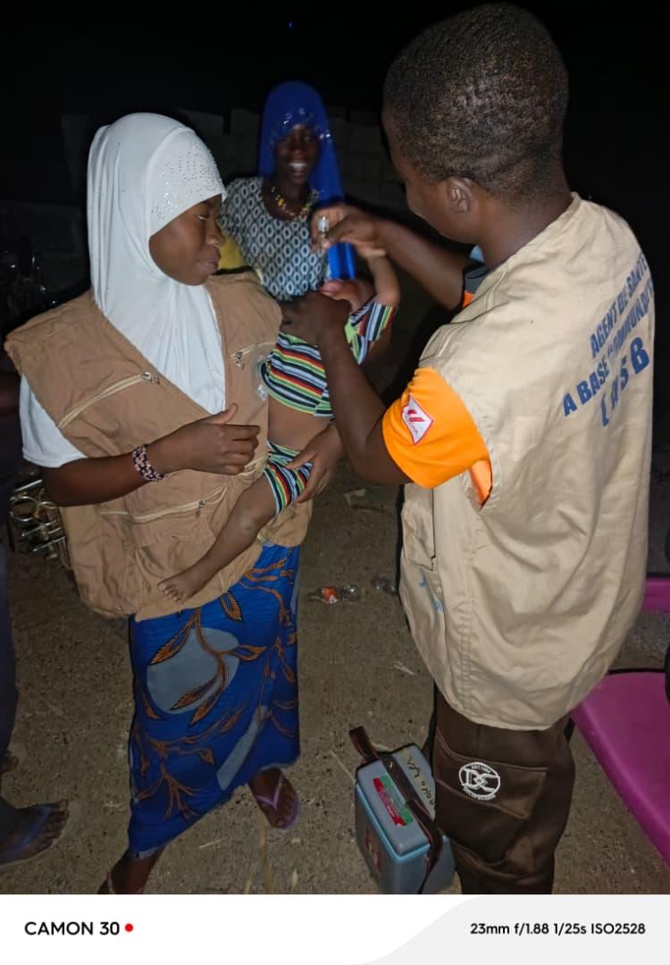 A nighttime vaccination scene showing a male health worker in a beige ASBC vest administering oral drops to a young child carried by a mother in a white hijab and beige vest. Another woman in blue looks on smiling. A vaccine cold box is visible on the ground.