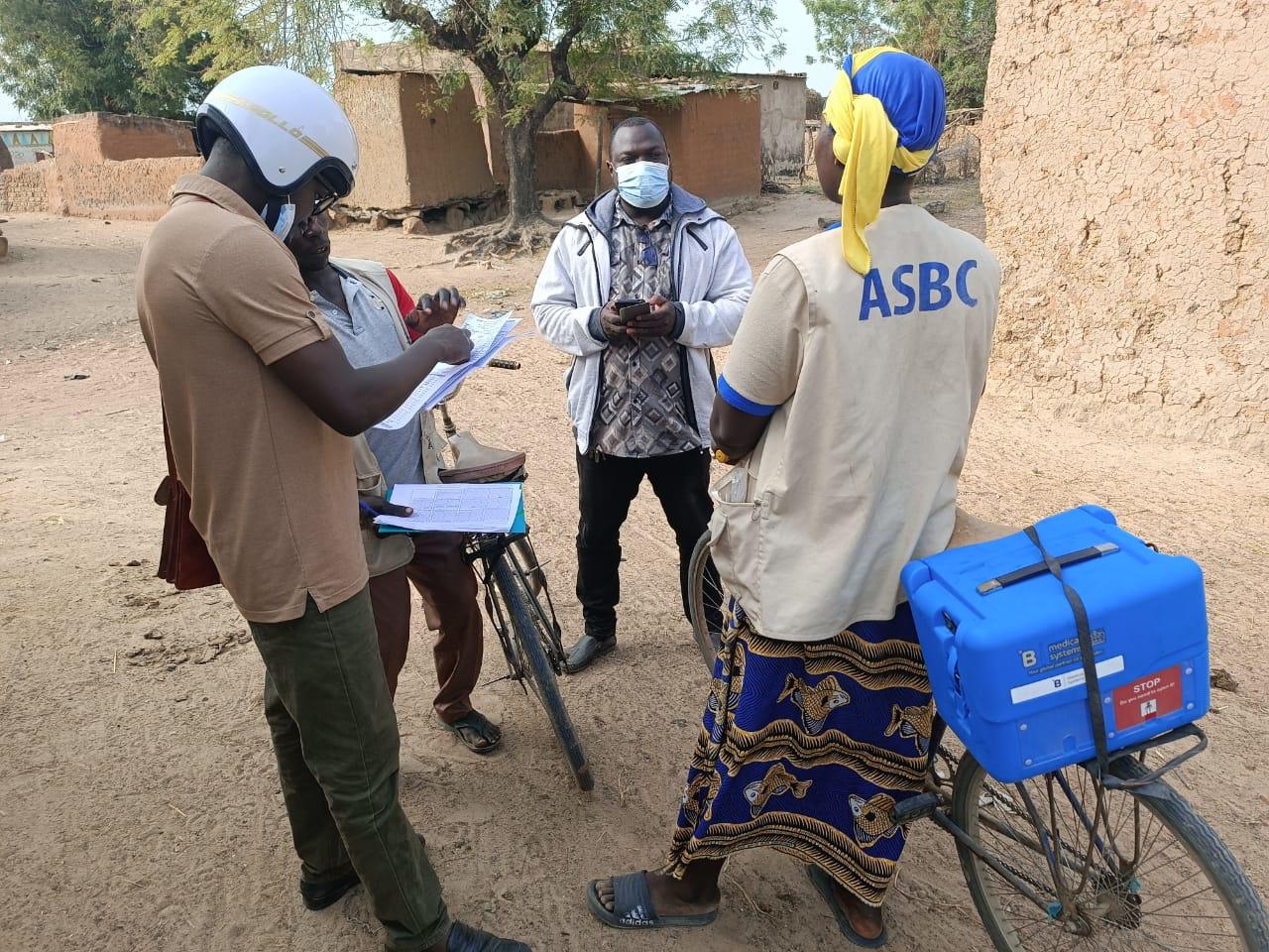 Community health workers discuss vaccination records during a field supervision visit in Nouna, Burkina Faso. A female ASBC worker wearing a yellow headscarf and vest marked "ASBC" stands with her bicycle carrying a blue UNICEF vaccine cold box. Three other health workers review paperwork beside her in a rural village setting with traditional mud houses in the background.