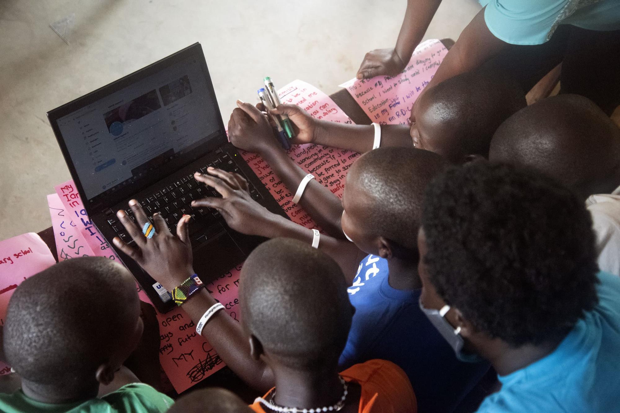 A group of children gather around a laptop, working together on a digital project, with handwritten notes and pens visible, highlighting youth engagement in digital education and teamwork.